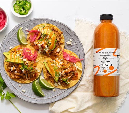 View A bottle of Terrapin Ridge Spicy Chipotle Squeeze sauce placed next to a gray plate of three loaded tacos with lime wedges, pickled red onions, avocado, and mango on a white background with a cream cloth.