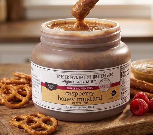 View A jar of Terrapin Ridge Farms Raspberry Mustard Pretzel Dip on a wooden surface, with pretzels, raspberries, and a spoonful of the dip shown around it.