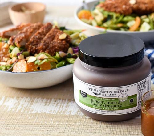 View A jar of Terrapin Ridge Farms Ginger Miso & Honey Dressing, shown in the foreground with bowls of prepared food in a kitchen setting.