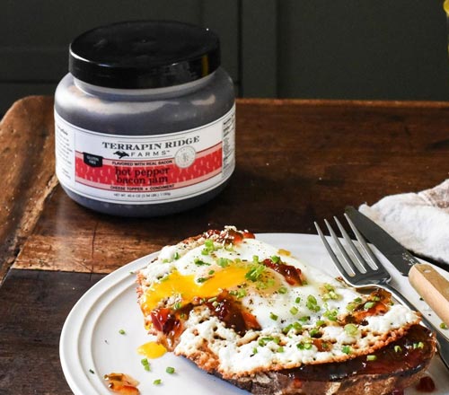 View A jar of Terrapin Ridge Farms Hot Pepper Bacon Jam sits on a rustic wooden table next to a plate of prepared food with a fried egg, garnished with chives and served with a fork and knife.