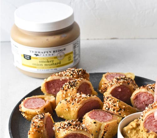 View Terrapin Ridge Farms Smokey Onion Mustard in a jar with a white lid, shown behind a black plate of sliced how dog rolls topped with sesame seeds, with a small bowl of mustard dip in the foreground.