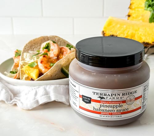 View A jar of Terrapin Ridge Farms Pineapple Habanero Sauce in the foreground, with a plate of shrimp tacos and pineapple slices blurred in the background on a light countertop.